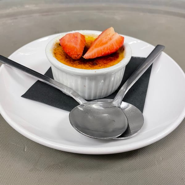 Crème brûlée in a white ramekin, topped with two strawberry halves, on a white plate with spoons.