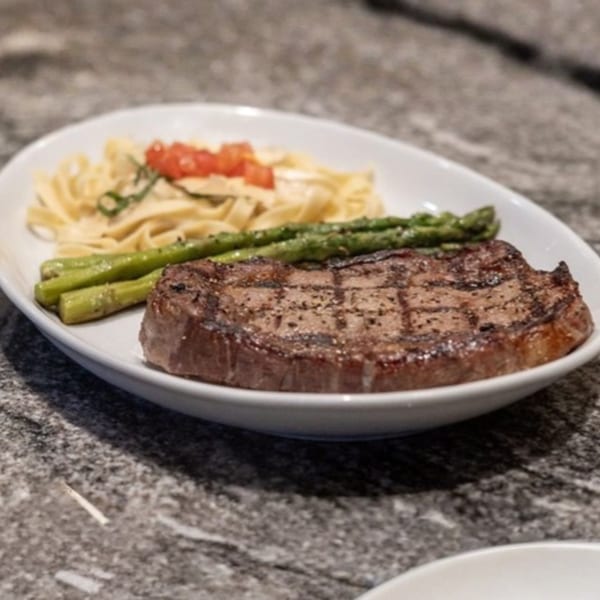 Grilled steak with asparagus and pasta, garnished with diced tomatoes and herbs, on a white plate.