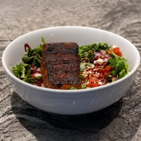 A white bowl filled with a dark seared salmon fillet on a bed of greens, tomatoes, and feta.