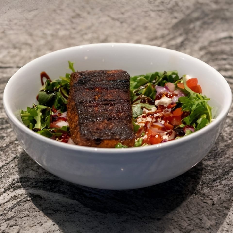 A white bowl filled with a dark seared salmon fillet on a bed of greens, tomatoes, and feta.