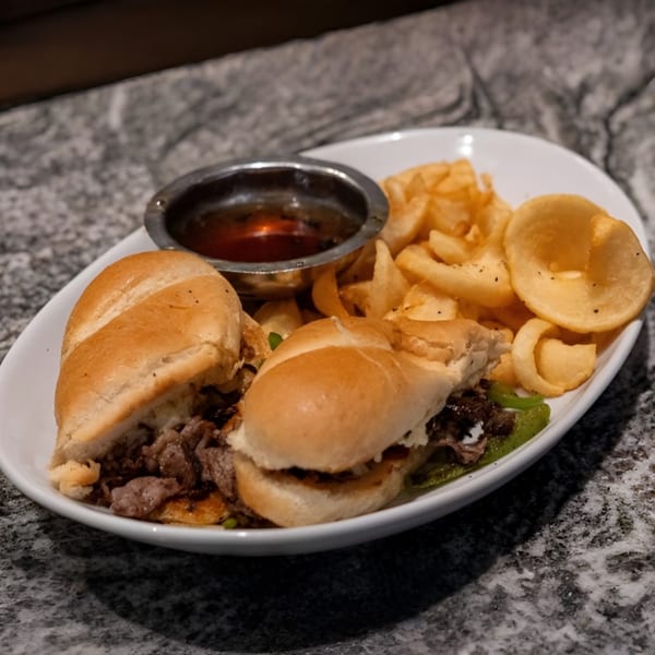 Two steak and cheese sliders with a side of crispy fries and a dipping sauce.