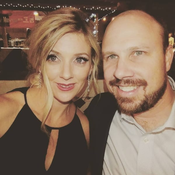 A smiling blonde woman and a bearded man pose for a selfie in a dimly lit restaurant.