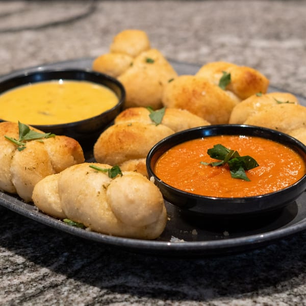 Garlic knots with two dipping sauces, one yellow and one red, on a dark plate.
