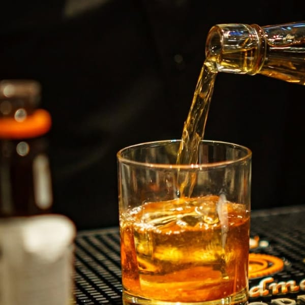 Whiskey pouring from a bottle into a glass with ice on a dark bar surface.