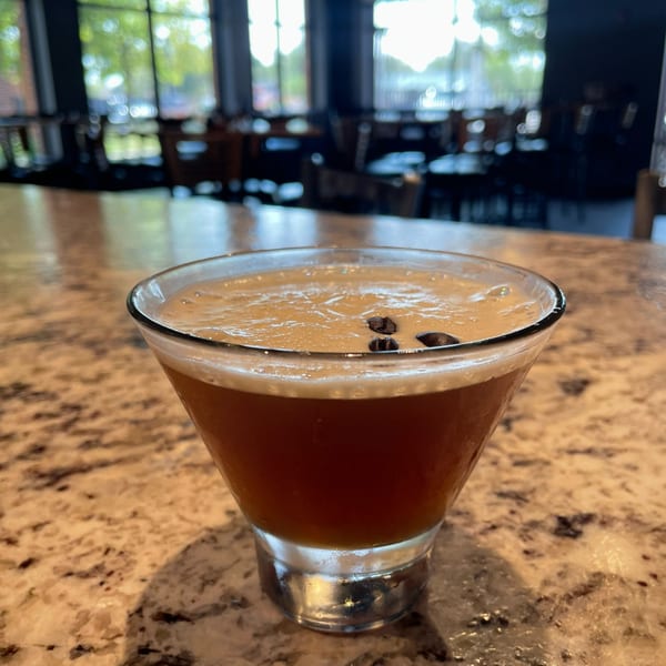 Espresso martini with coffee beans on top, on a speckled bar counter with a restaurant in the background.